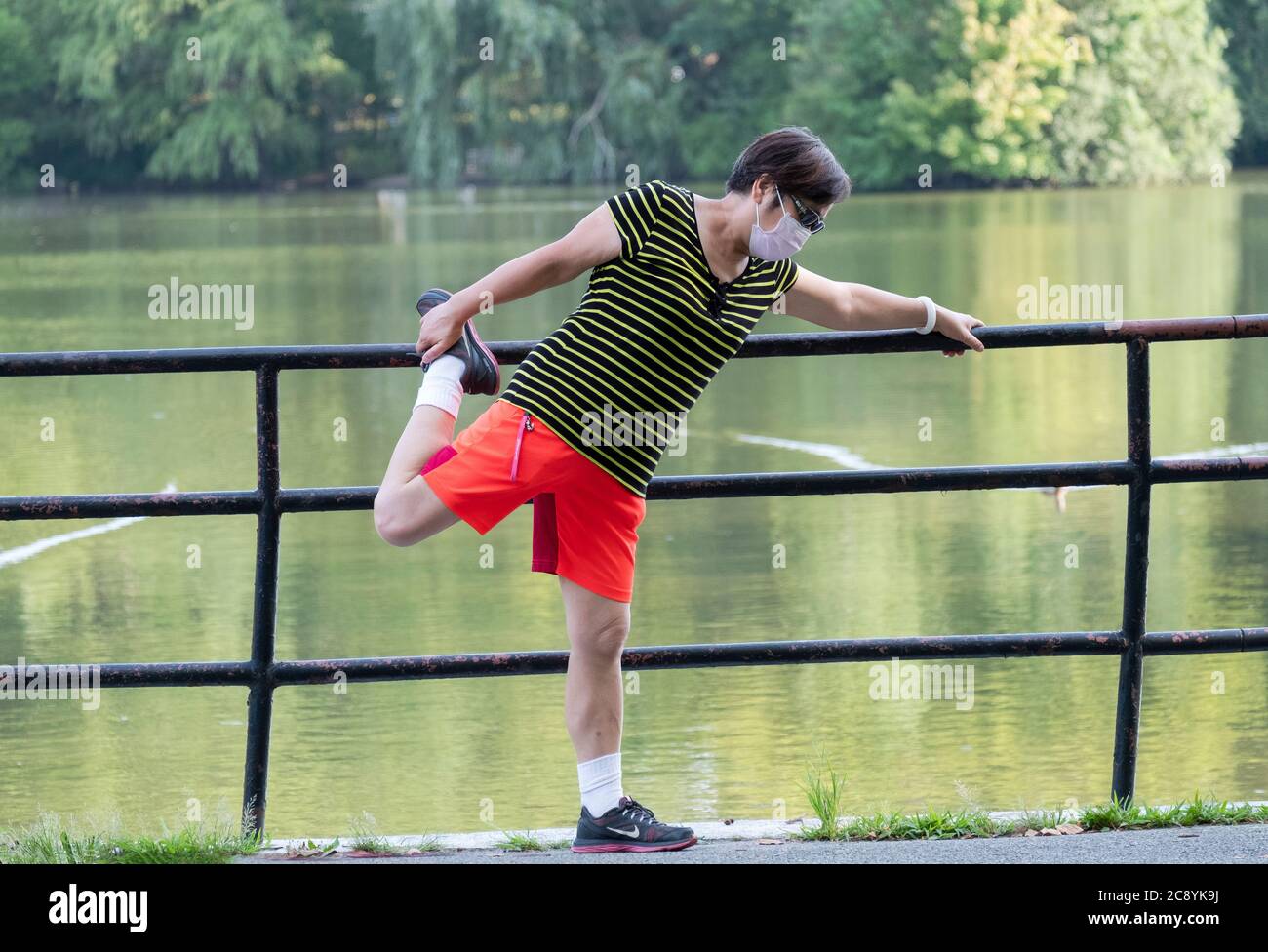 Eine asiatisch-amerikanische Frau, die eine chirurgische Maske trägt, dehnt ihre Quadricep-Muskeln vor dem Training. In Kissena Park, Flushing, New York City. Stockfoto