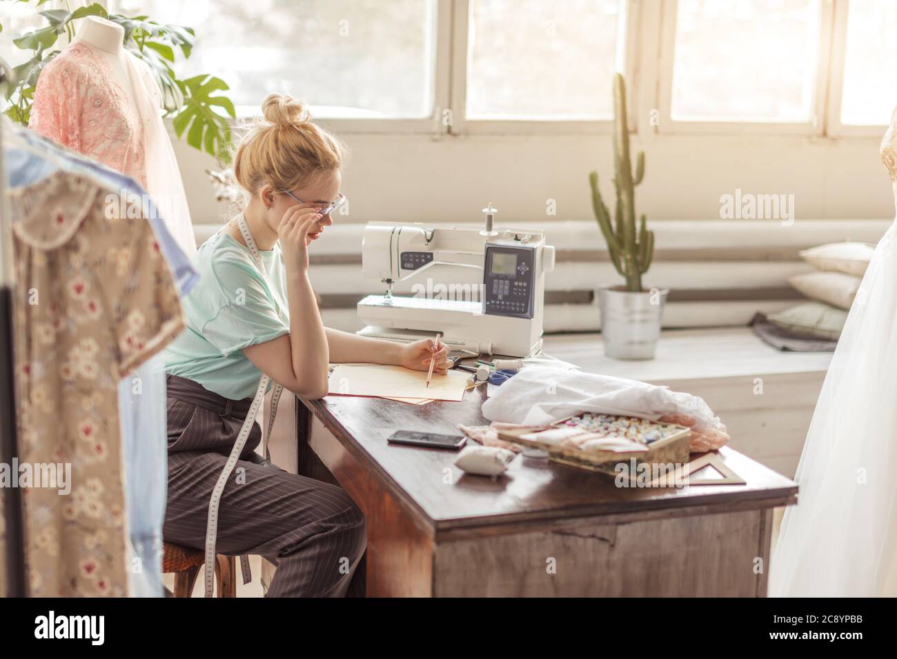 Attraktive junge Couturier in Brillen sitzen auf dem Desktop mit Skizzen, in die Kamera schauen und lächeln, während der Arbeit im Atelier im alten Stil Stockfoto