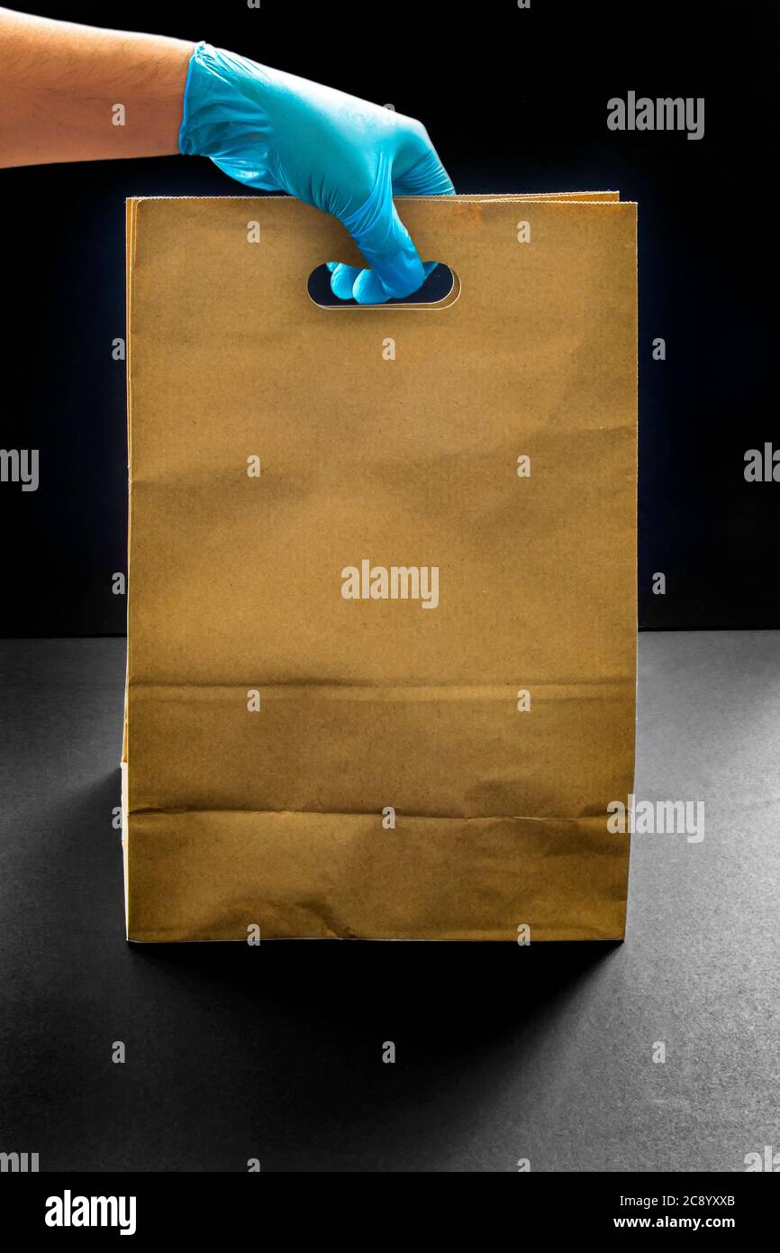 Eine Person mit Handschuhen, die eine braune Einkaufstasche auf dunklem Hintergrund hält. Persönliche Schutzausrüstung Einkaufskonzept. Stockfoto