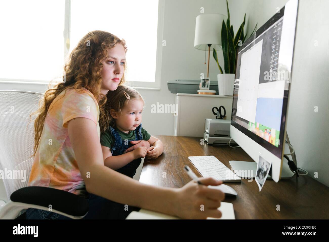 Junge Mutter mit ihrer Tochter auf dem Schoß, die von zu Hause aus arbeitet Am Schreibtisch Stockfoto