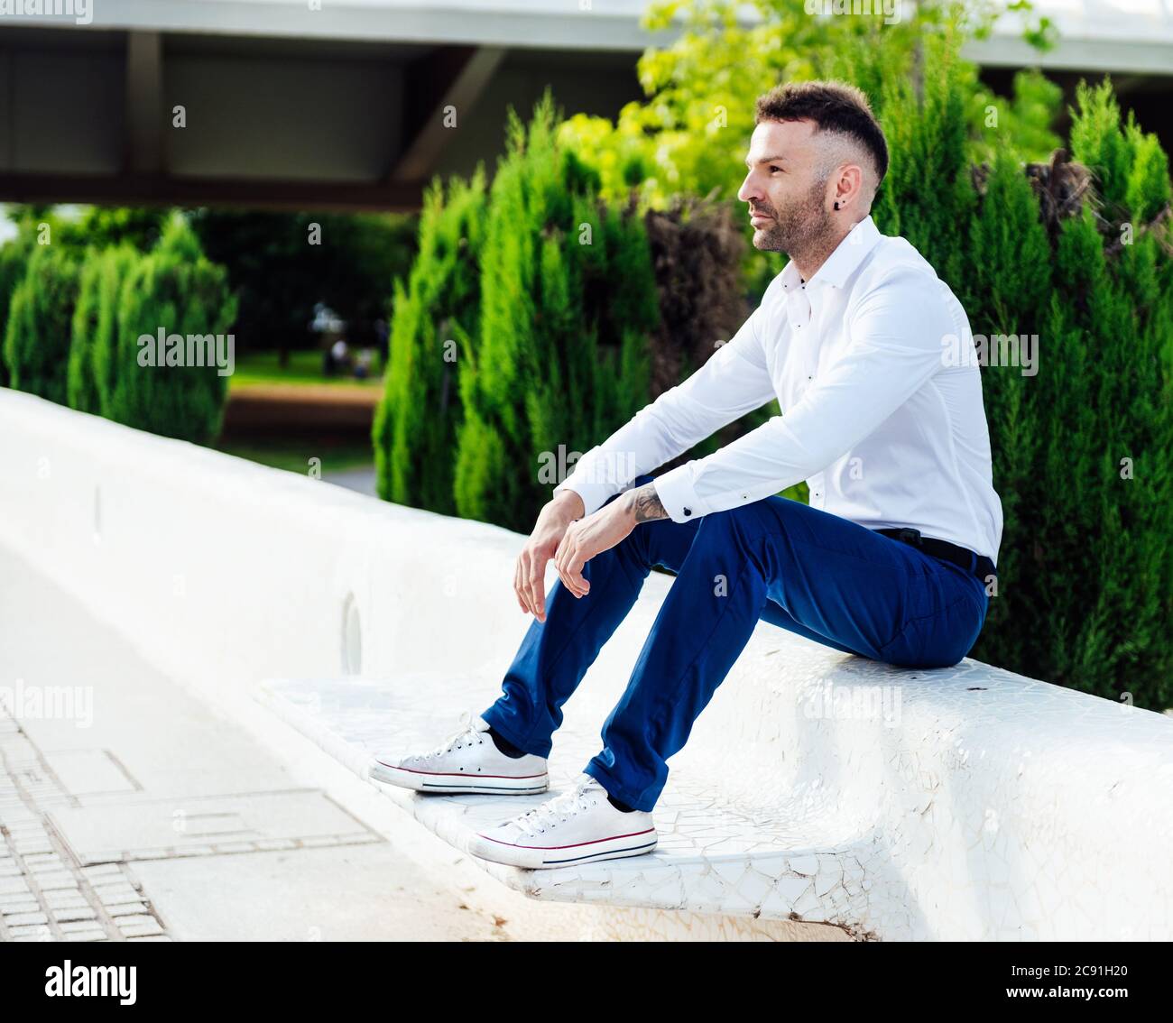 Junger Mann in weißem Hemd, blauer Hose und Ohrring sitzt auf einer Bank  und schaut zur Seite Stockfotografie - Alamy