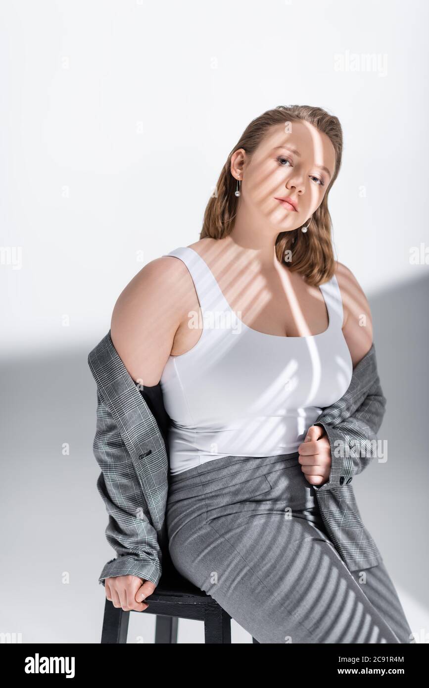 Attraktive, selbstbewusste plus size Mädchen, die Blazer aus, während auf Stuhl auf weiß im Sonnenlicht mit Schatten sitzen Stockfoto