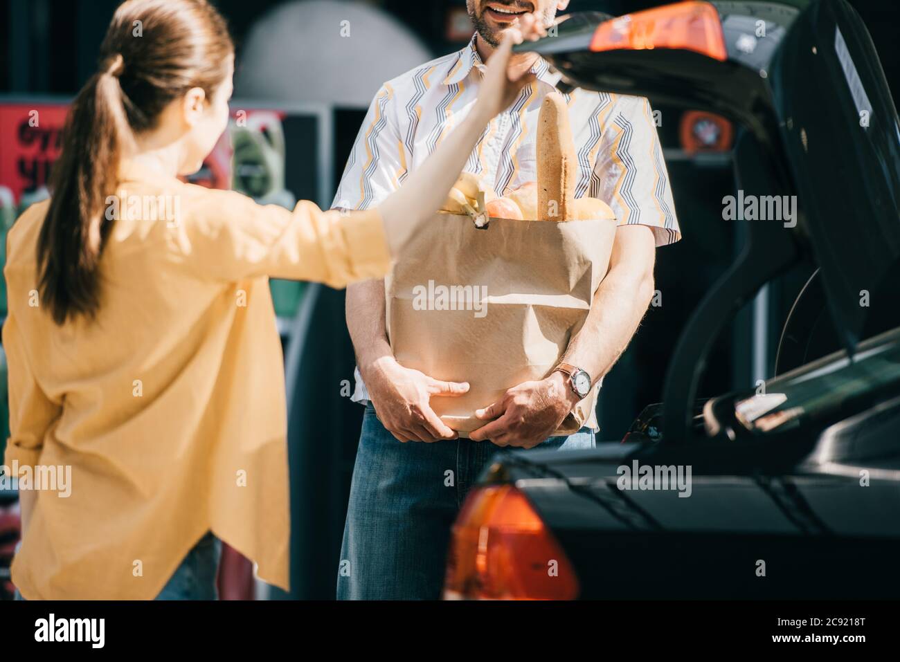 Selektiver Fokus auf lächelnden Mann, der Einkaufstasche mit Essen in der Nähe von Frau und Kofferraum auf der städtischen Straße hält Stockfoto