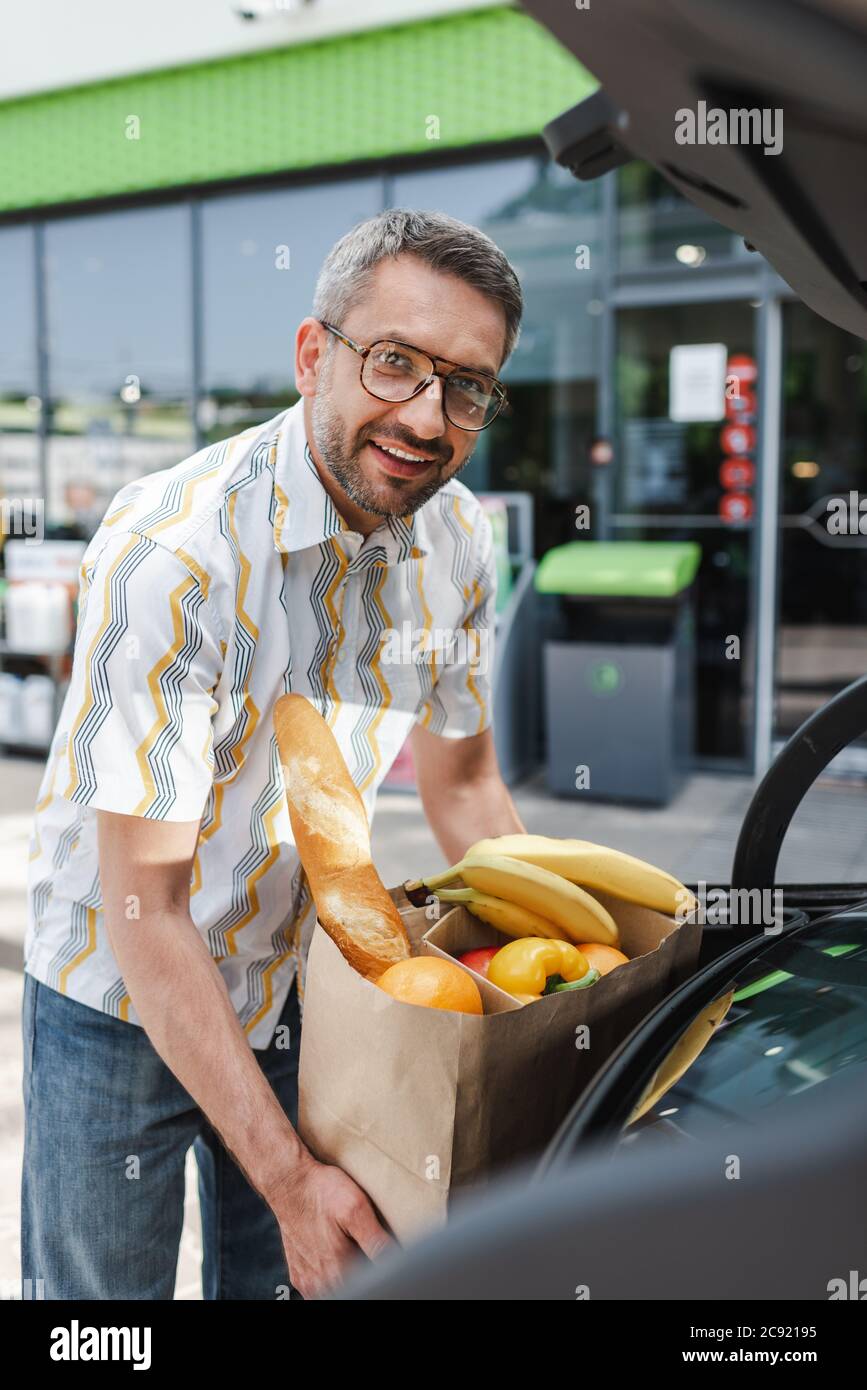 Selektiver Fokus des lächelnden Mannes, der Einkaufstasche mit Nahrung in offenen Kofferraum auf der städtischen Straße setzt Stockfoto