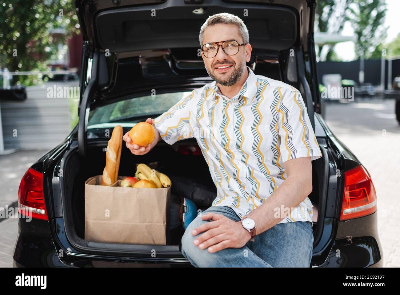 Lächelnder Mann, der Orange in der Nähe einer Einkaufstasche mit Essen und offenem Kofferraum auf der Stadtstraße hält Stockfoto