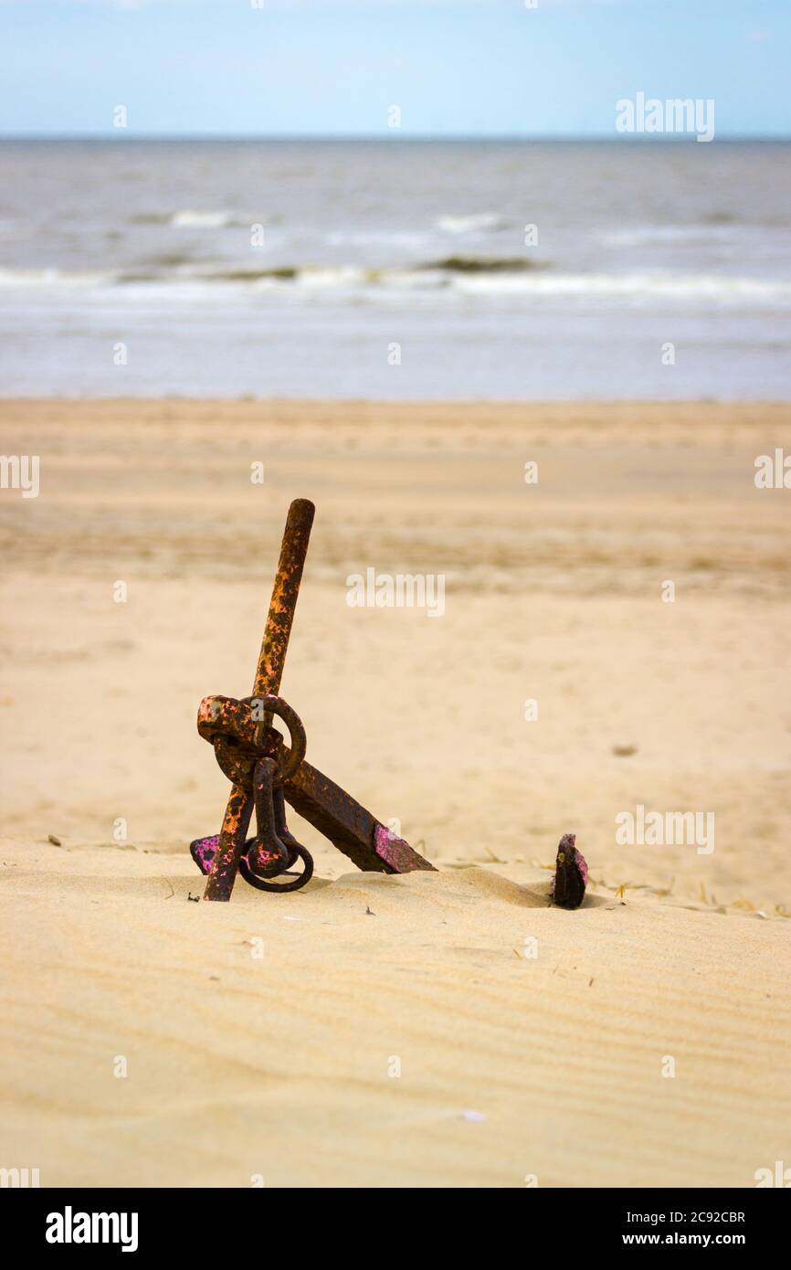 Rostige orange und rosa Anker liegen am Strand, teilweise versunken in Sand vor kleinen Meereswellen. Ruhige Landschaft, ruhig, leicht rau. Stockfoto