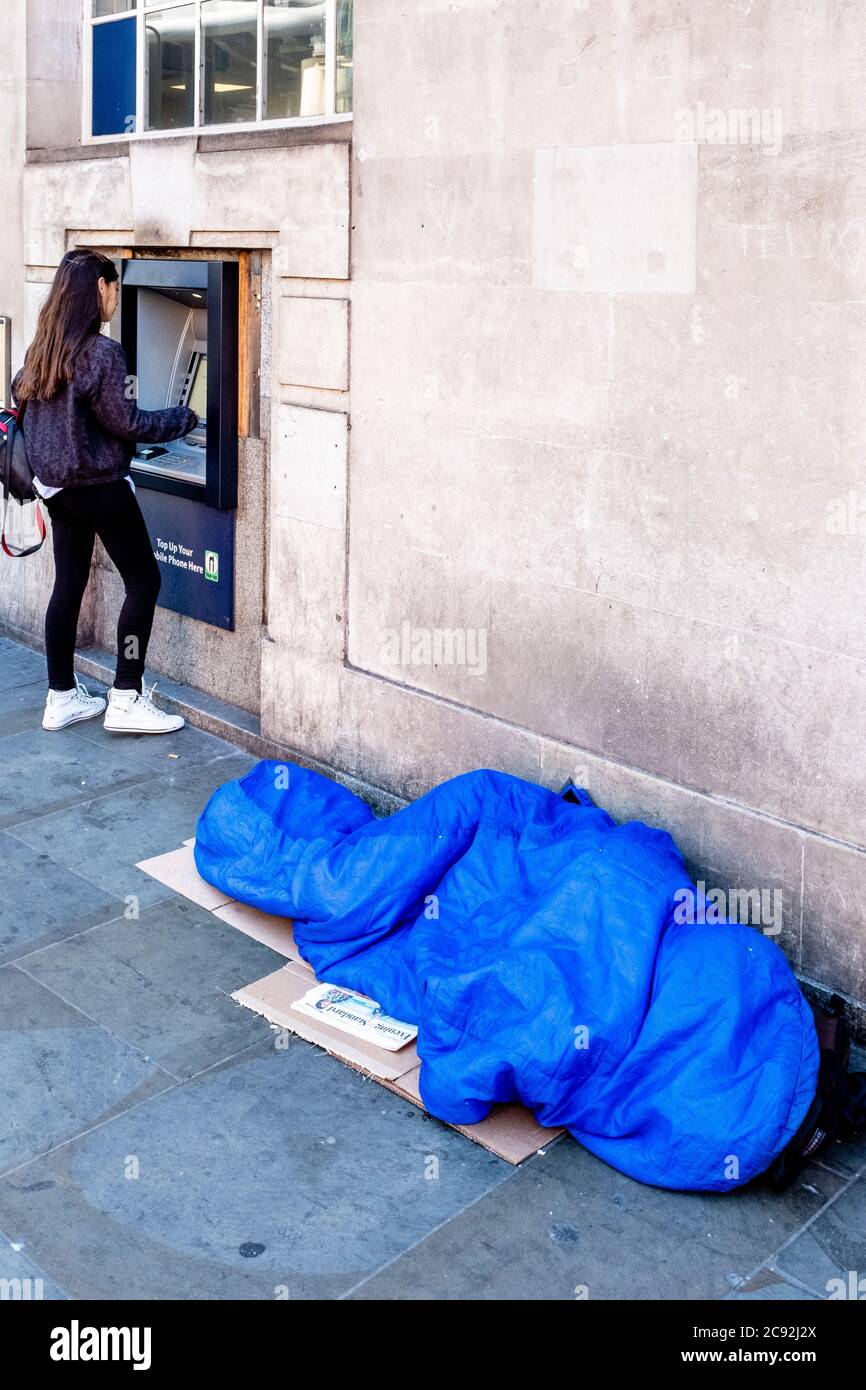 Eine obdachlose Person schläft in EINEM Schlafsack, während EINE junge Frau Geld an einem Geldautomaten in der Borough High Street, London, England abzieht. Stockfoto