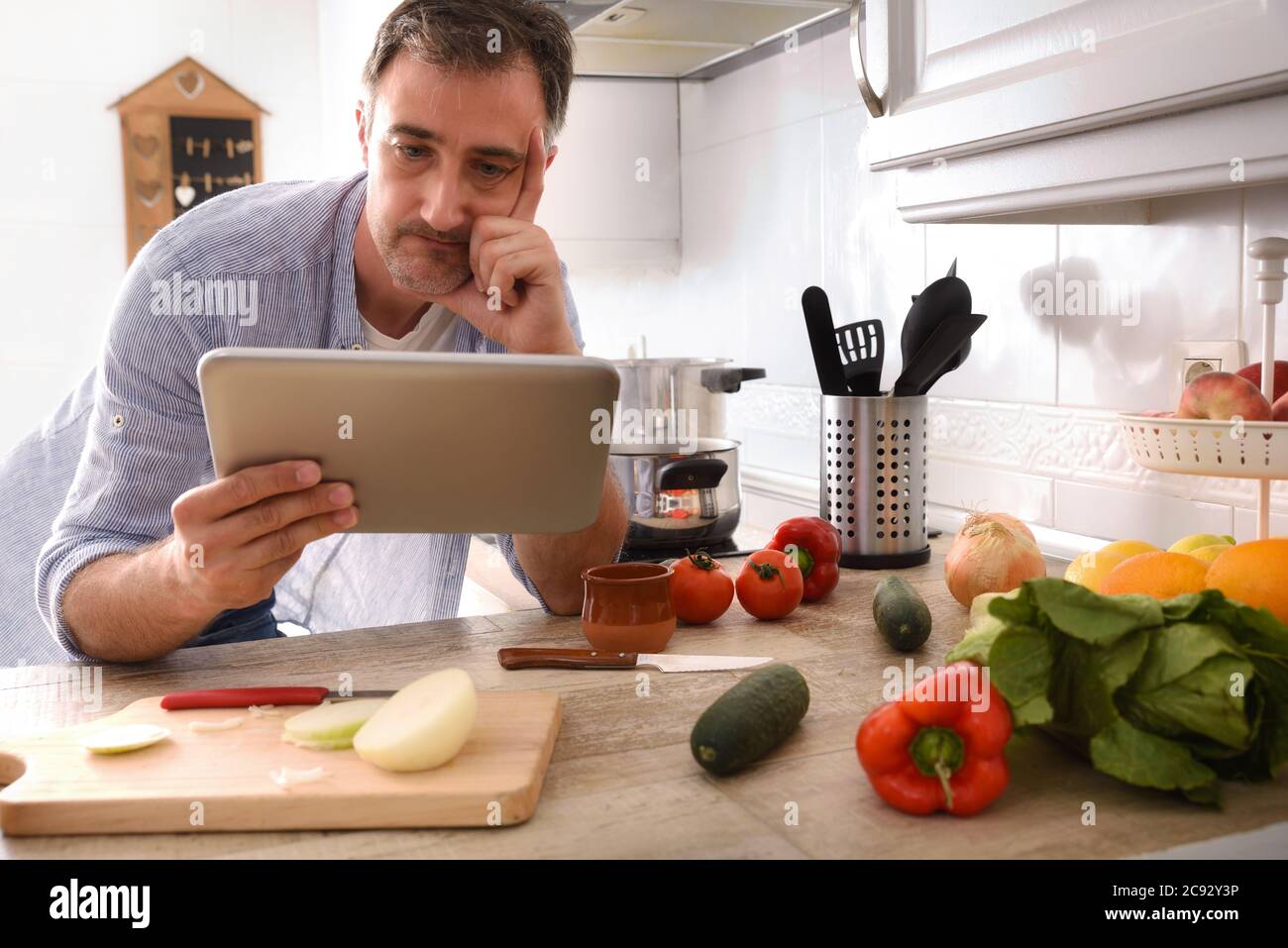 Zweifelhafter Mann, der zu Hause kocht und dabei das Rezept auf einer Tablette ansieht, die mit der Hand auf dem Gesicht auf der Küchenbank liegt Stockfoto