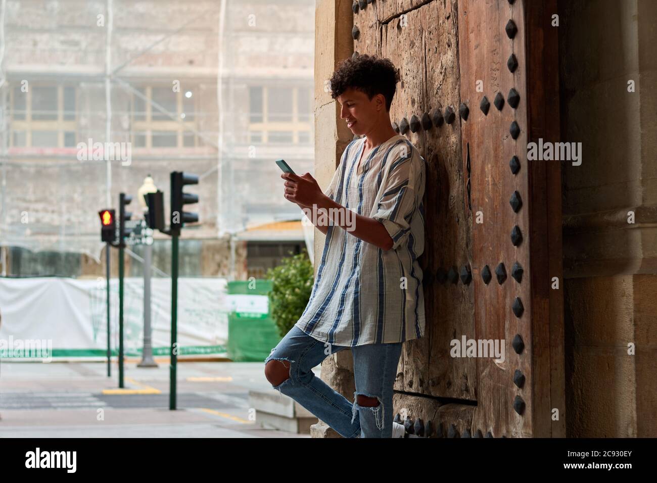 Der junge Afro lehnt sich an ein hölzernes Tor und schaut auf seine Telefon Stockfoto