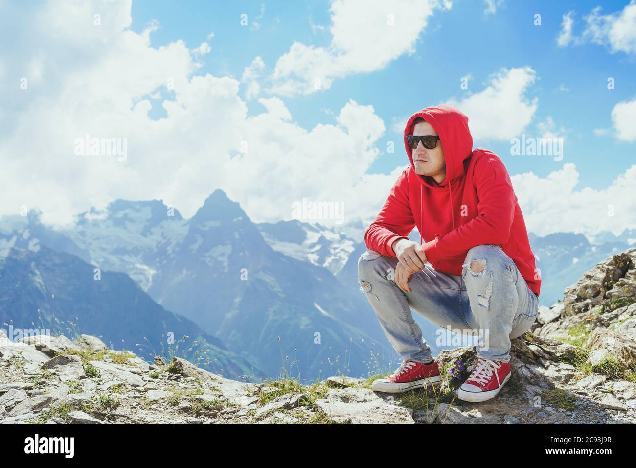 Junger Mann in Sonnenbrille sitzt auf dem Berg bei sonnigem Wetter. Erwachsener Rüde in rotem Kapuzenpullover mit Kapuze und schöner Aussicht in der bergigen Gegend. Stockfoto