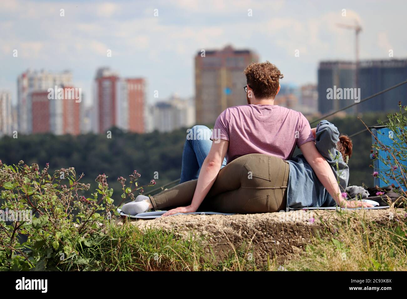 Verliebtes Paar, das auf einem Hügel auf einem Stadthintergrund sitzt. Sommer Freizeit und Entspannung, Konzept des Träumens und Zukunftsplanung, romantische Datum Stockfoto