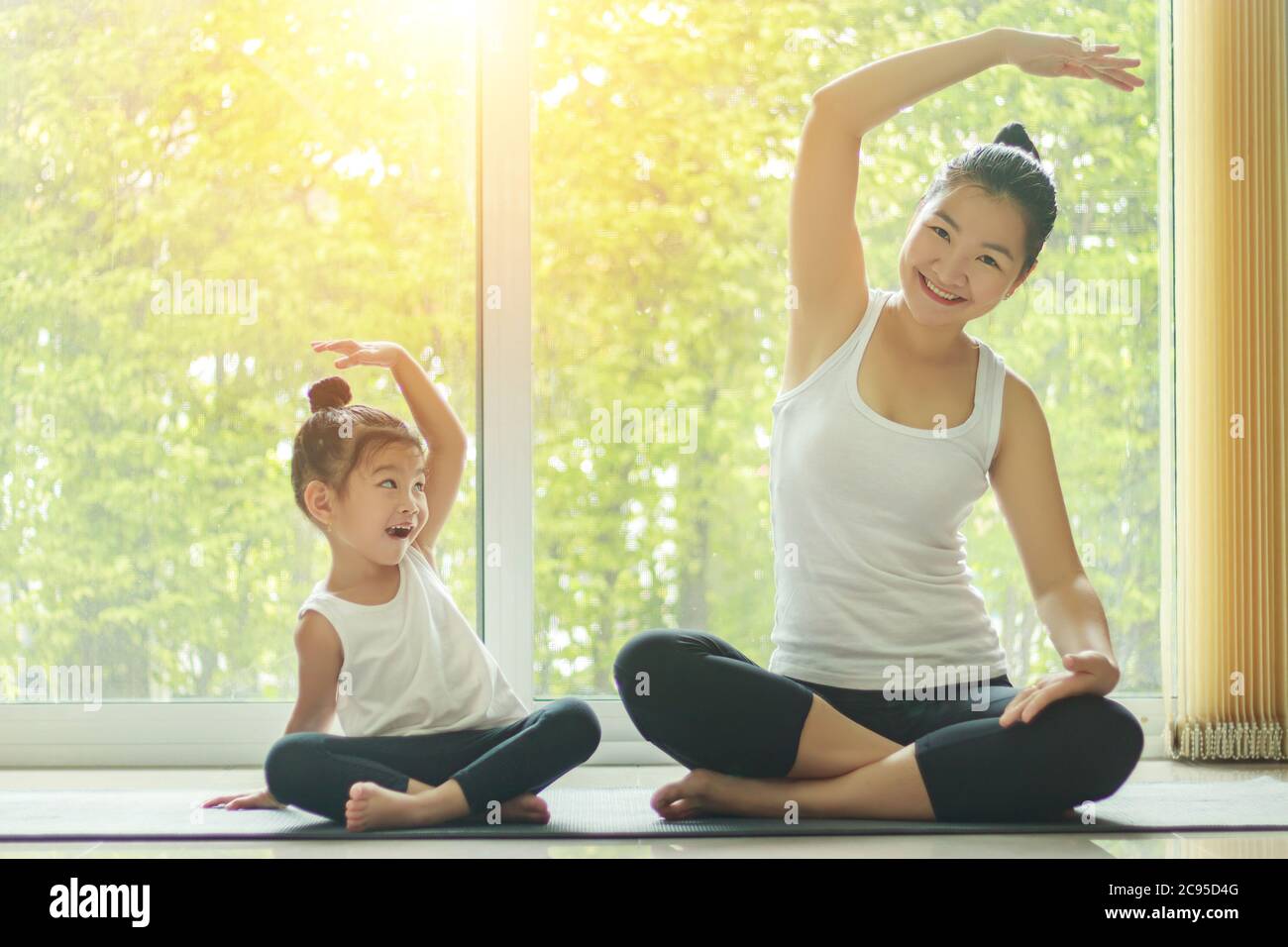 Asiatische Mama Yoga zu Hause mit einer entzückenden Tochter neben ihr sitzen, versuchen, die Haltung der Mutter mit einem lächelnden Gesicht zu imitieren Stockfoto