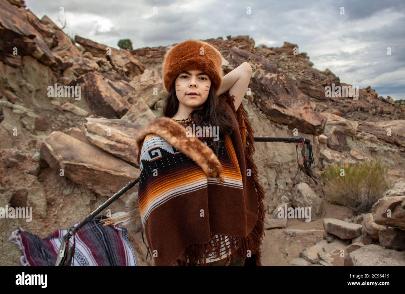 Junge hispanische Frau mit Pfeil und Bogen und als Trapper in Poncho und Pelzhut in der ojito Wilderness Area in New Mexico, USA Stockfoto