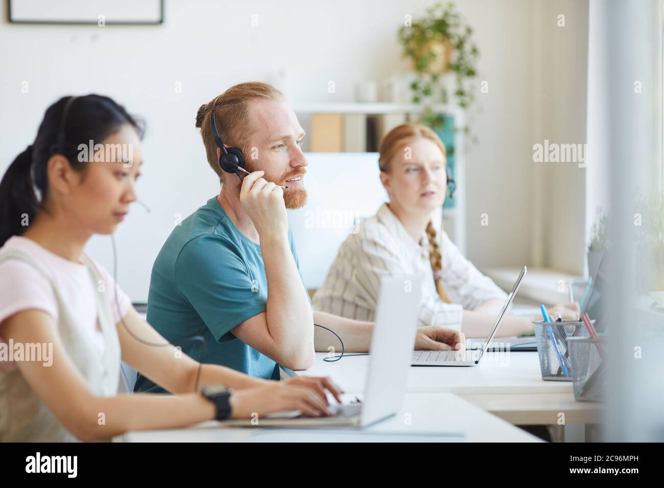 Gruppe von Personen, die im Callcenter arbeiten, die Kopfhörer tragen, sitzen am Tisch und beraten Kunden Stockfoto