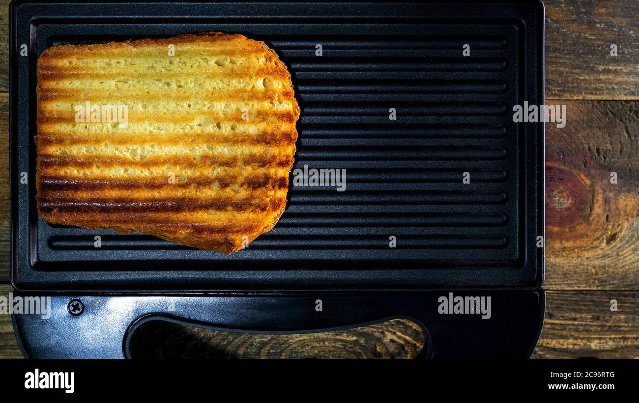 Gegrilltes weißes Toastbrot mit goldener Kruste in Streifen liegt in offener schwarzer Sandwich-Maker, auf Holz rustikalen Tisch. Schnelles Kochen zu Hause. Draufsicht. Flat la Stockfoto