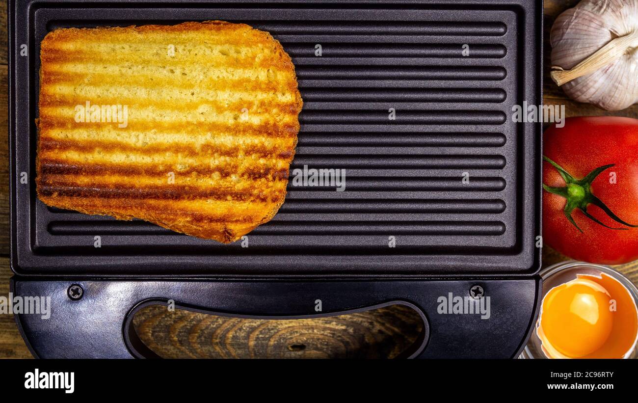Gegrilltes weißes Toastbrot mit goldener Kruste in Streifen liegt in offener schwarzer Sandwich-Maker, auf Holz rustikalen Tisch. Eigelb, Tomate, Knoblauch. Schnell zu Hause Stockfoto