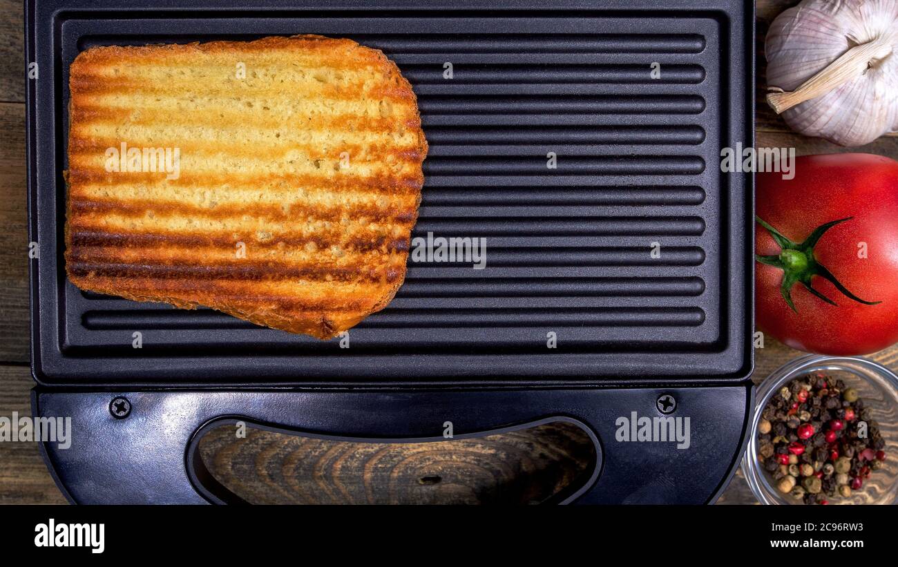 Gegrilltes weißes Toastbrot mit goldener Kruste in Streifen liegt in offener schwarzer Sandwich-Maker, auf Holz rustikalen Tisch. Gewürze, Tomaten, Knoblauch. Schnelles CO Stockfoto