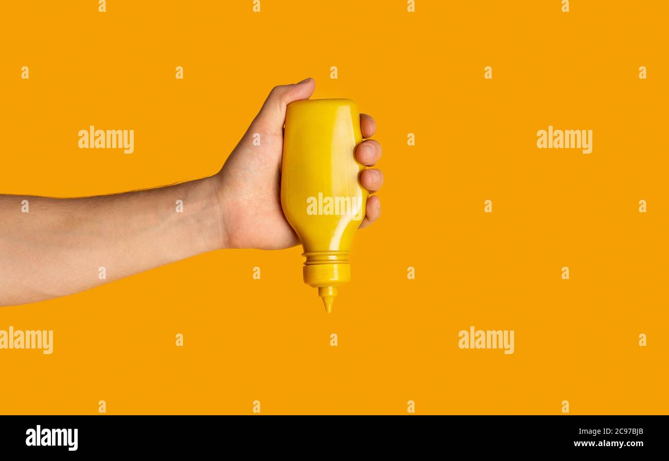 Männliche Hand drückt Flasche mit Senf auf orangenen Hintergrund, Nahaufnahme. Leerzeichen für Text Stockfoto
