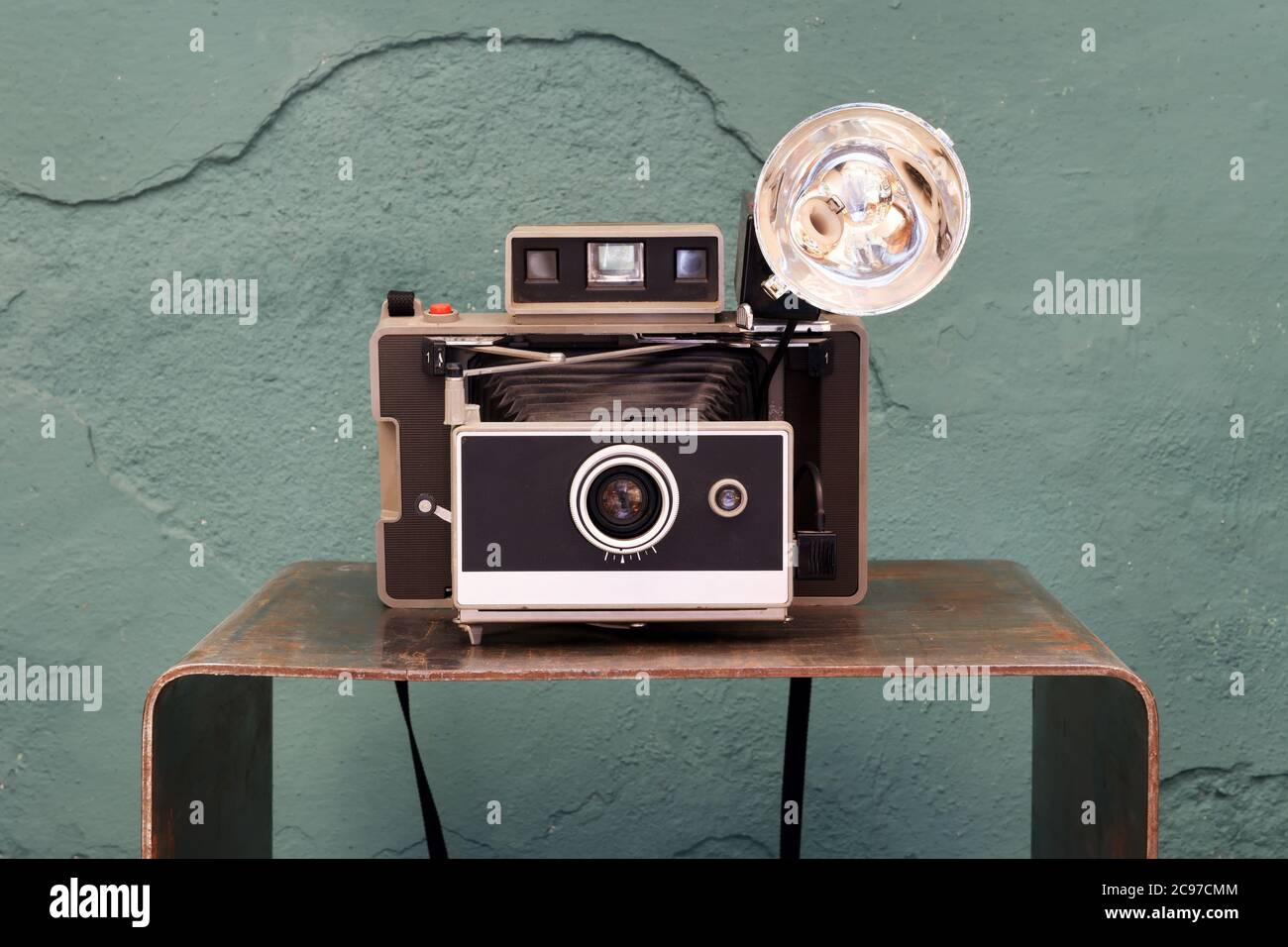 Alte Vintage-Sofortkamera mit Faltenbalg und Lampe auf einem kleinen Regal vor einer beschädigten grünen Strukturwand in einem fotografischen Konzept Stockfoto