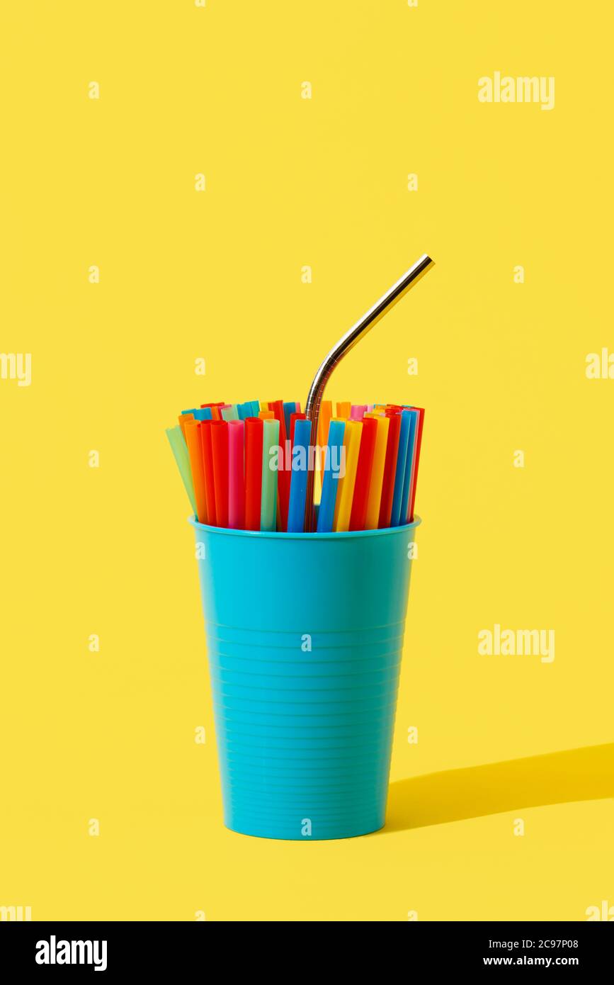 Ein wiederverwendbarer Trinkhalm aus Edelstahl, der sich aus einigen Einweg-Trinkhalmen aus Kunststoff in verschiedenen Farben in einer blauen Vase auf einer gelben BA herausstellt Stockfoto