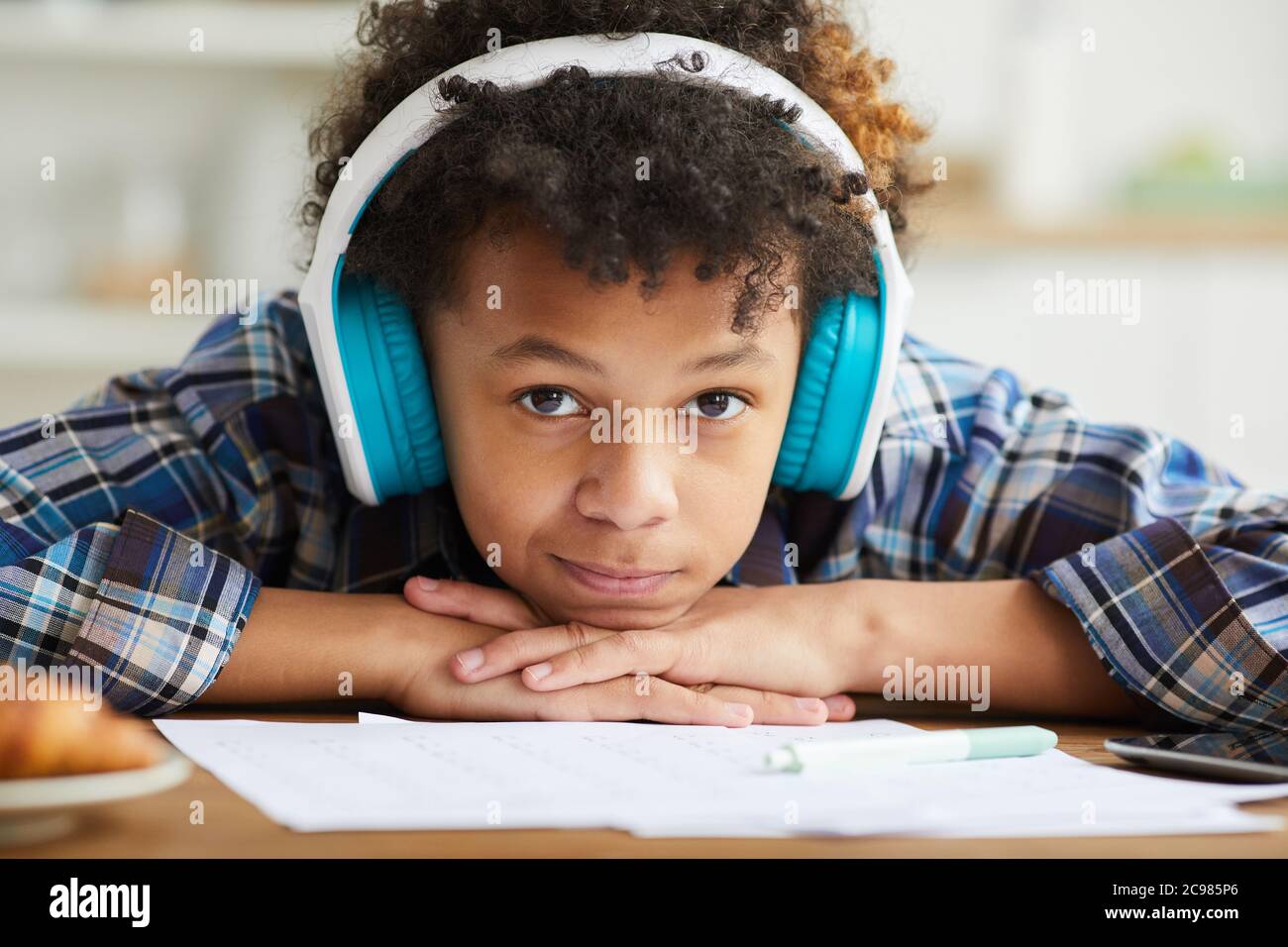 Nahaufnahme eines afrikanischen Schuljungen mit lockigen Haaren in Kopfhörern, der die Kamera anschaut, während er am Tisch sitzt und studiert Stockfoto