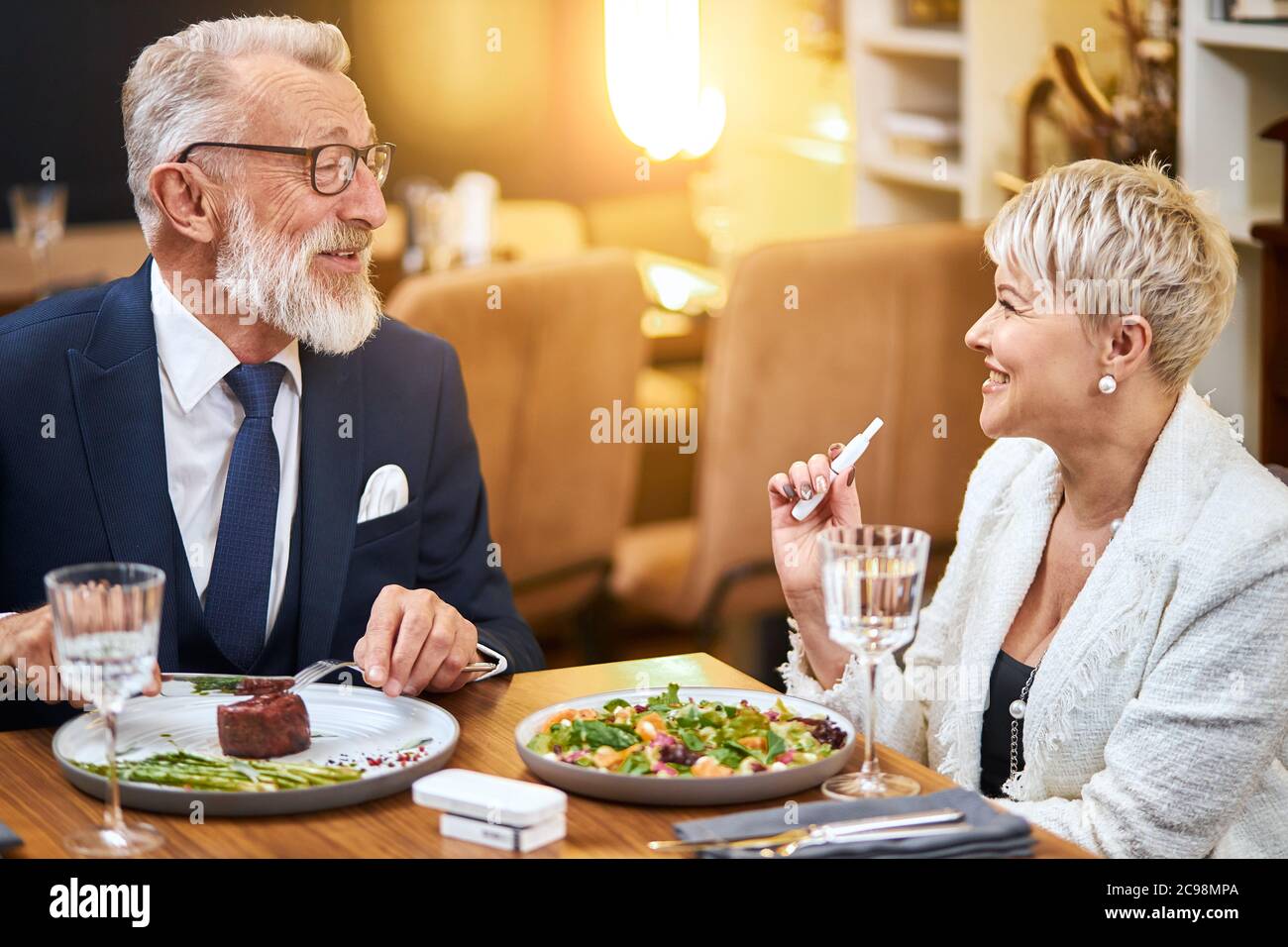 ltere Paare essen im reichen Restaurant zu Abend, whrend sie IQOS, E- Zigarette, benutzen. Schner Mann im Smoking lcheln, Frau ansehen und  sprechen Stockfotografie - Alamy