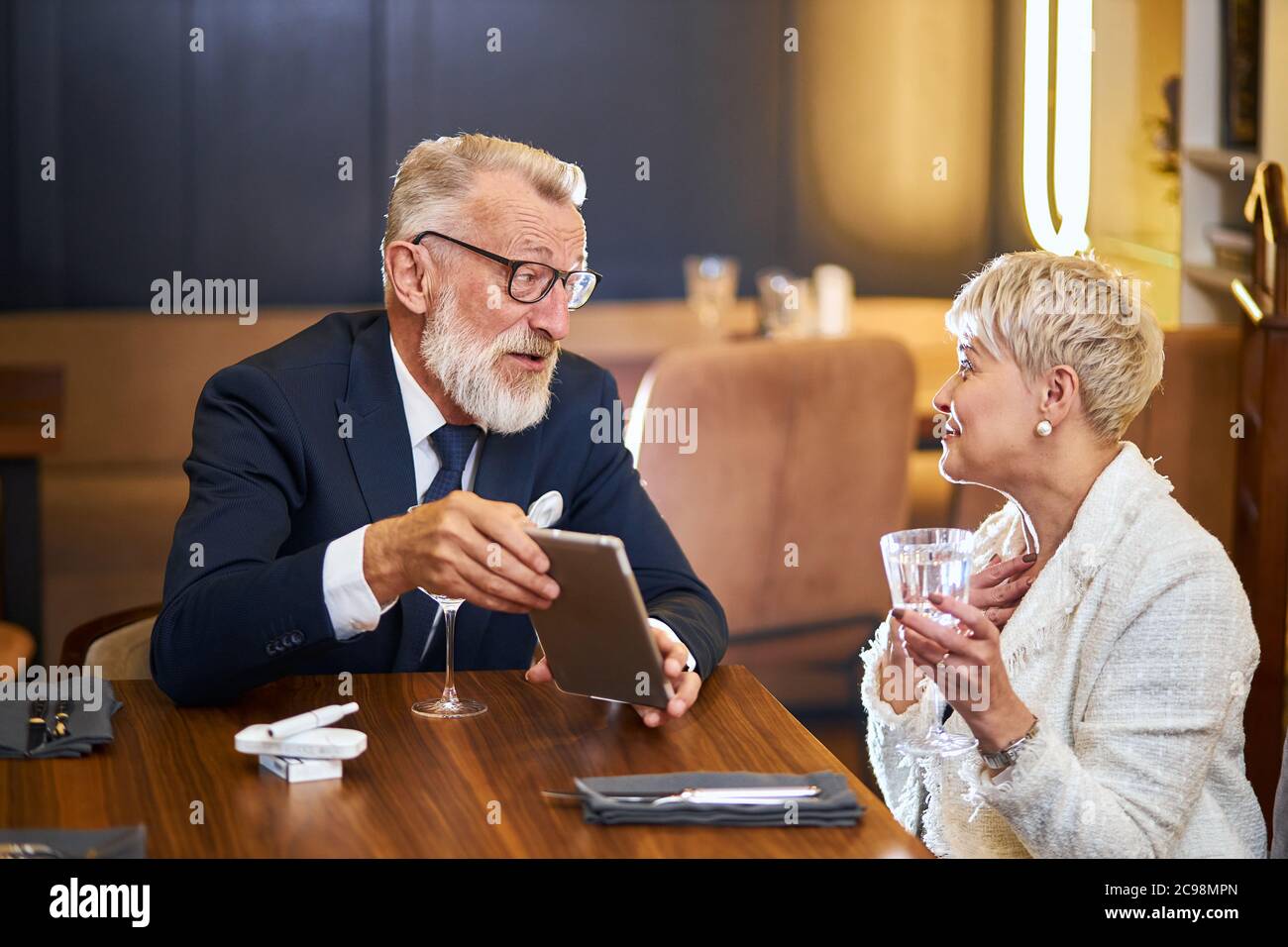 Attraktive Senioren verwenden moderne Technologien, Tablet im Restaurant. Grauhaariger Mann in Smoking, Brille und Frau in weißem Blazer Stockfoto