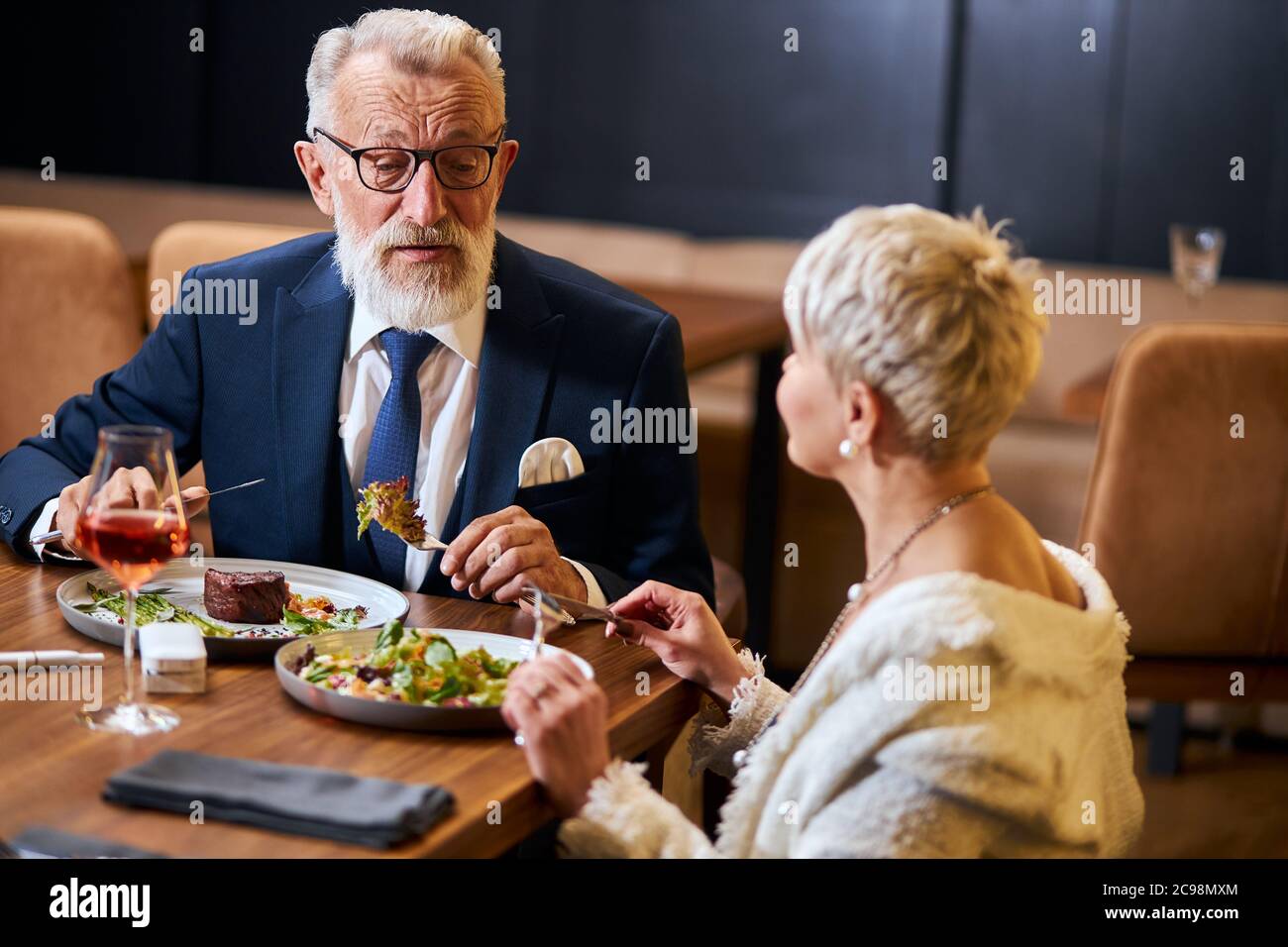 Hübsche Dame und älterer grauhaariger Mann im Smoking, der sich im Restaurant freundlich unternahm. Kollegen nach der Arbeit diskutieren und essen Stockfoto