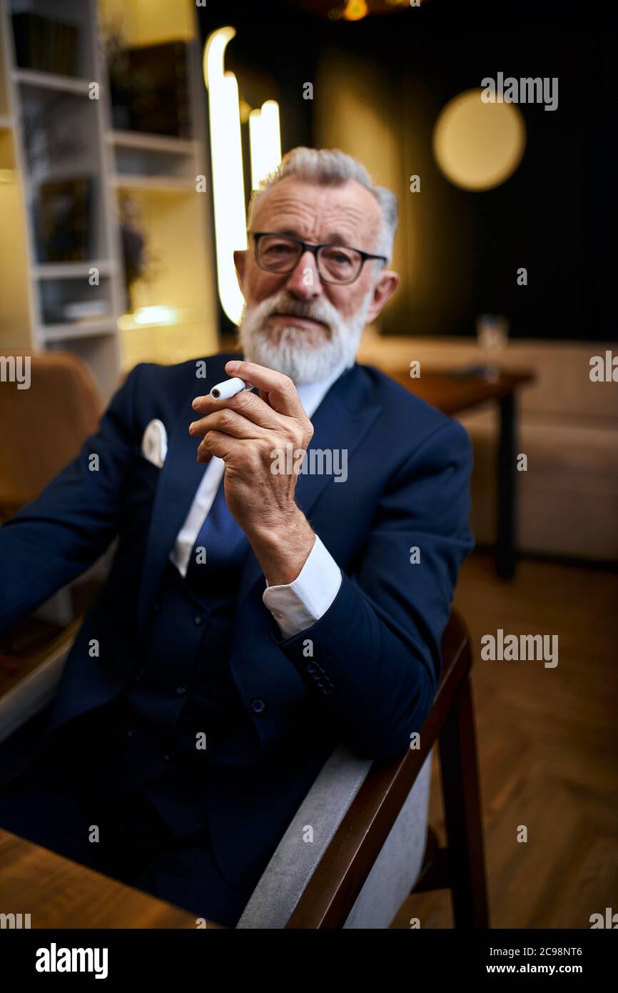 Schöner grauhaariger älterer Mann in Gläsern in Anzug Rauch IQOS im Restaurant, erkunden Zigarette halten in den Händen Stockfoto