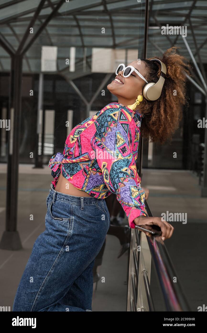 Seitenansicht einer positiven afroamerikanischen Dame in legerer Kleidung, die im Headset Musik hört, während sie auf dem Geländer in der Nähe des Gebäudes gelehnt steht Stockfoto