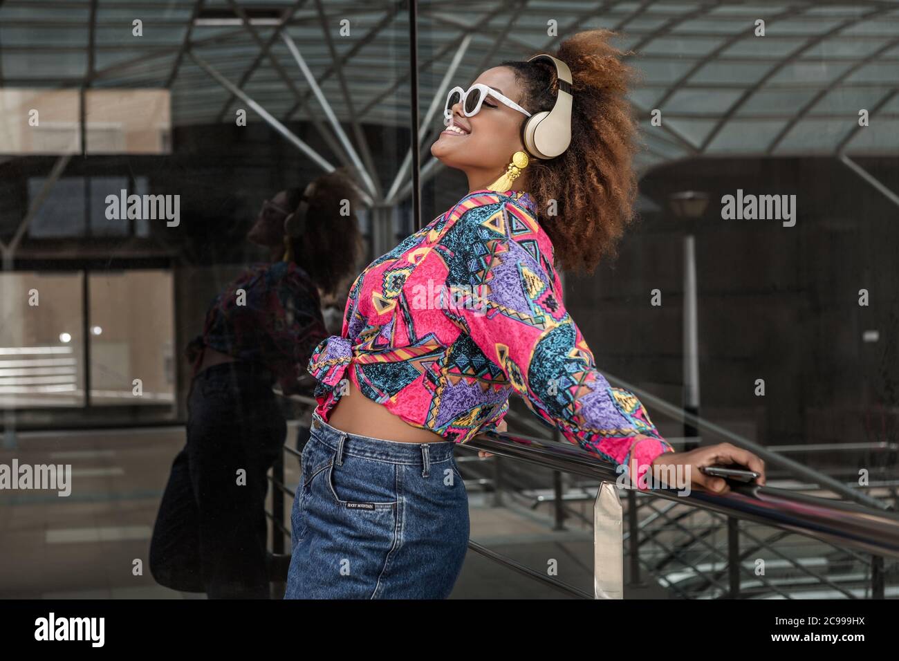 Seitenansicht des Inhalts afroamerikanische Dame in Jeans und Sonnenbrille, die Musik im schnurlosen Headset genießt, während sie mit dem Smartphone auf dem Handrai steht Stockfoto