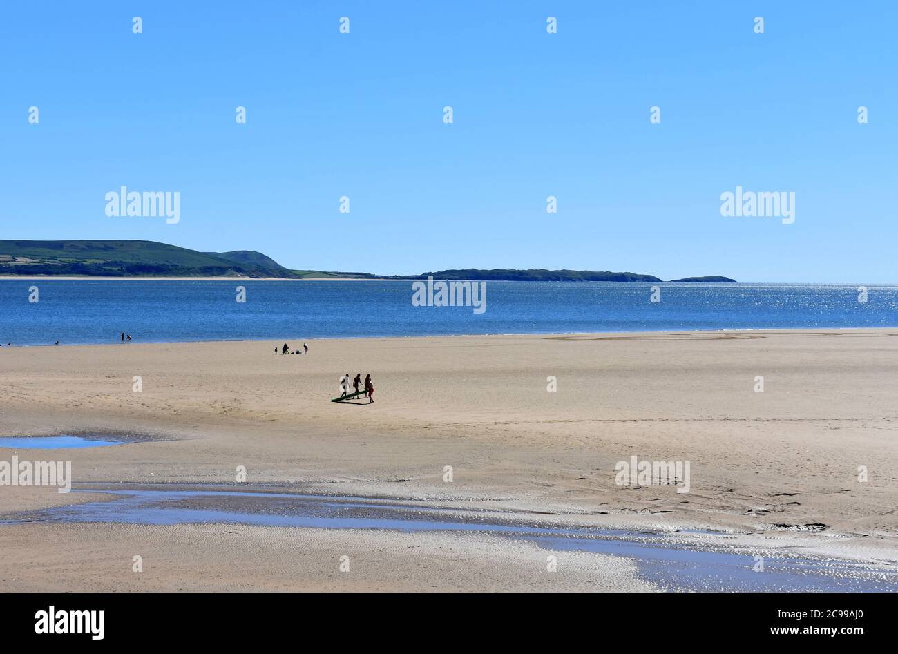 Urlauber, die bei Ebbe ein Surfbrett am Strand von Burry Port West tragen, Burry Port, Carmarthenshire, Wales Stockfoto