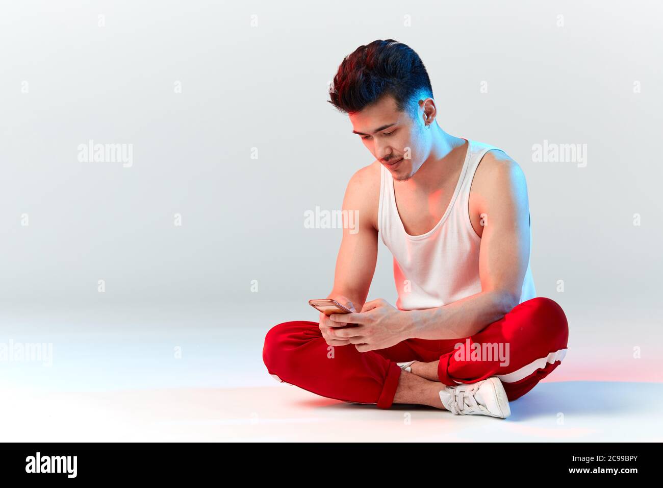 Junge sportliche stilvolle männliche Tänzerin in roten Jogginghosen Überprüfung seines Smartphones im Studio über weißem Hintergrund. Asiatische Hipster bboy lächelnd bei Kamera befo Stockfoto
