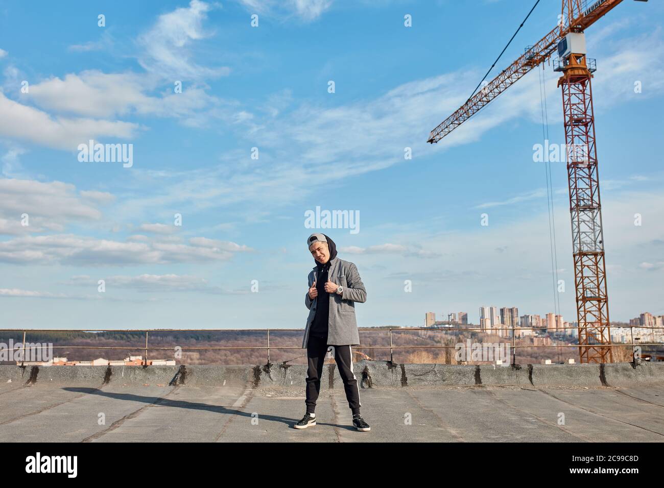 Asiatische Hip-Hop Mann Tänzerin in schwarzen Sporthosen gekleidet, Sweatshirt posiert draußen auf dem Dach über blauen Himmel mit Bau Kran auf Hintergrund Stockfoto
