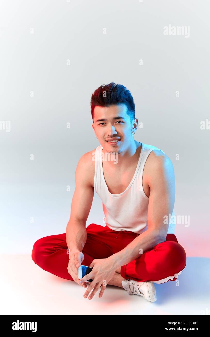 Junge sportliche stilvolle männliche Tänzerin in roten Jogginghosen posiert mit gekreuzten Leggs isoliert im Studio auf weißem Hintergrund. Asiatische Hipster bboy lächelnd an c Stockfoto