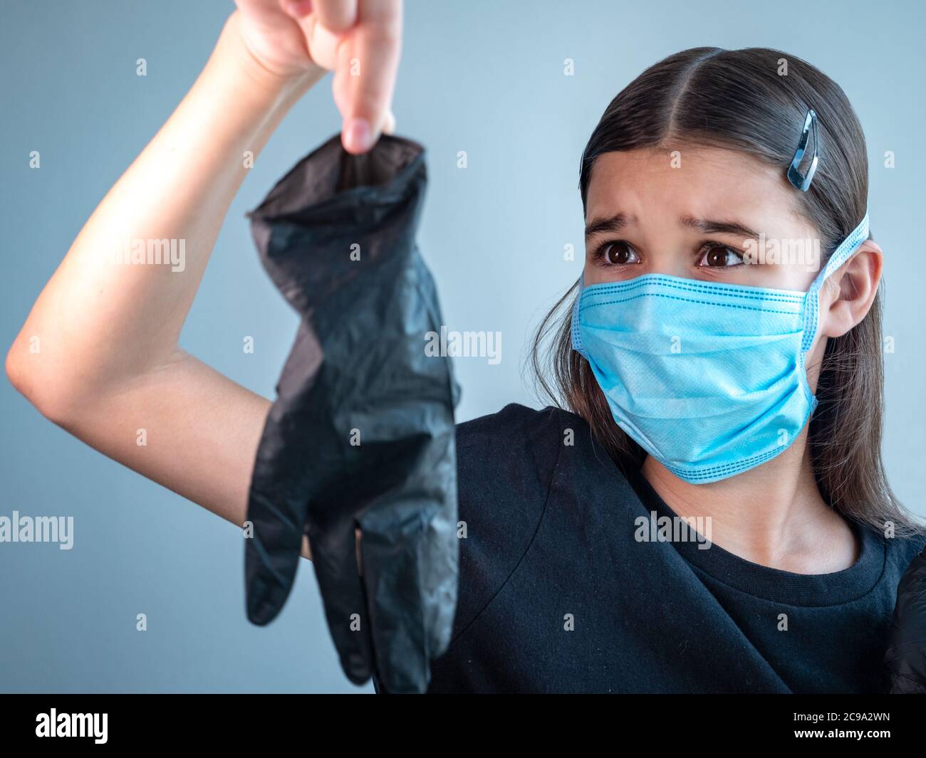 Verängstigte weibliche Teenager-Mädchen in einer blauen schützenden medizinischen Gesichtsmaske hält einen schwarzen Latex-Handschuh durch Spitzen ihrer Finger auf Armlänge. Coronavirus Stockfoto