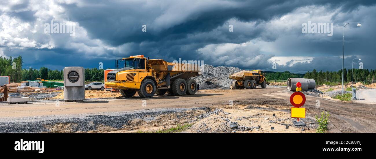 Panoramablick auf zwei schwere industrielle gelbe Muldenkipper in der Nähe der Baustelle mit bestückten Baustoffen geparkt, dunkelblaue Wolken vor ra Stockfoto