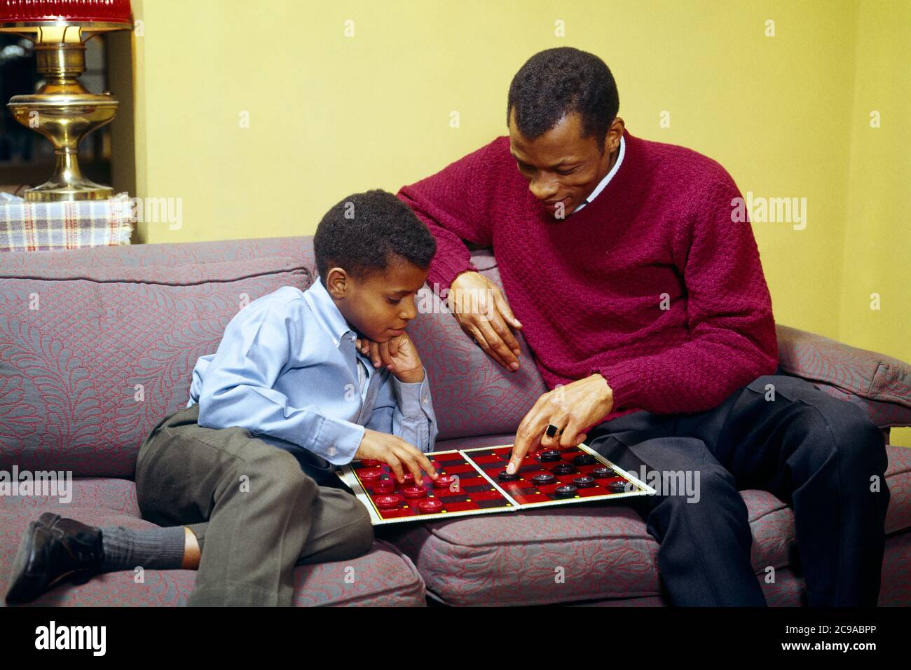 1960S 1970S AFRICAN-AMERICAN VATER UND SOHN SITZEN AUF COUCH SPIELEN DAME SPIEL VATER HILFT, ZEIGT AUF EINE MÖGLICHE BEWEGUNG - KJ5656 PHT001 HARS PAAR FARBE ALT ZEIT LEHRE NOSTALGIE ALTE MODE 1 JUGENDLICH COUCH STIL KOMMUNIKATION WETTBEWERB STARKE SÖHNE FAMILIEN FREUDE LIFESTYLE ELTERNSCHAFT ZU HAUSE LEBEN KOPIEREN RAUM FREUNDSCHAFT HALBE LÄNGE PERSONEN FÜRSORGLICHE MÄNNER VÄTER CHECKERS BEWEGEN GLÜCK HIGH-ANGLE-STRATEGIE AFROAMERIKANER AFROAMERIKANER UND VÄTER SLACKS SCHWARZE ETHNIZITÄT STOLZ POTENZIELLE VERBINDUNG PFLEGER PERSÖNLICHE BINDUNG ZUNEIGUNG BRETT SPIEL ZUSAMMENARBEIT EMOTION JUGENDLICHE MITTE ERWACHSENE Stockfoto