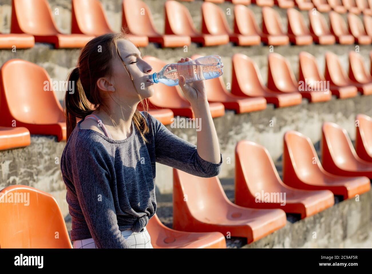 Sportliche junge attraktive Mädchen in Sportbekleidung entspannen nach hartem Training sitzen und trinken Wasser aus speziellen Sportflasche nach dem Laufen auf einem Stadion Stockfoto