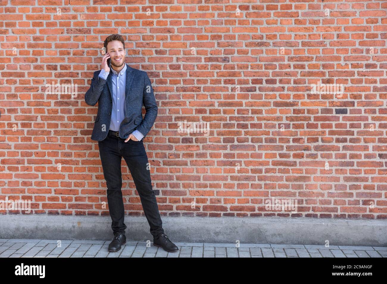 Mann sprechen Geschäft am Telefon hält Smartphone in der Stadt Straße in smart Casual tragen stehen vor Backsteinmauer städtischen Hintergrund. Glücklicher Kaukasier Stockfoto