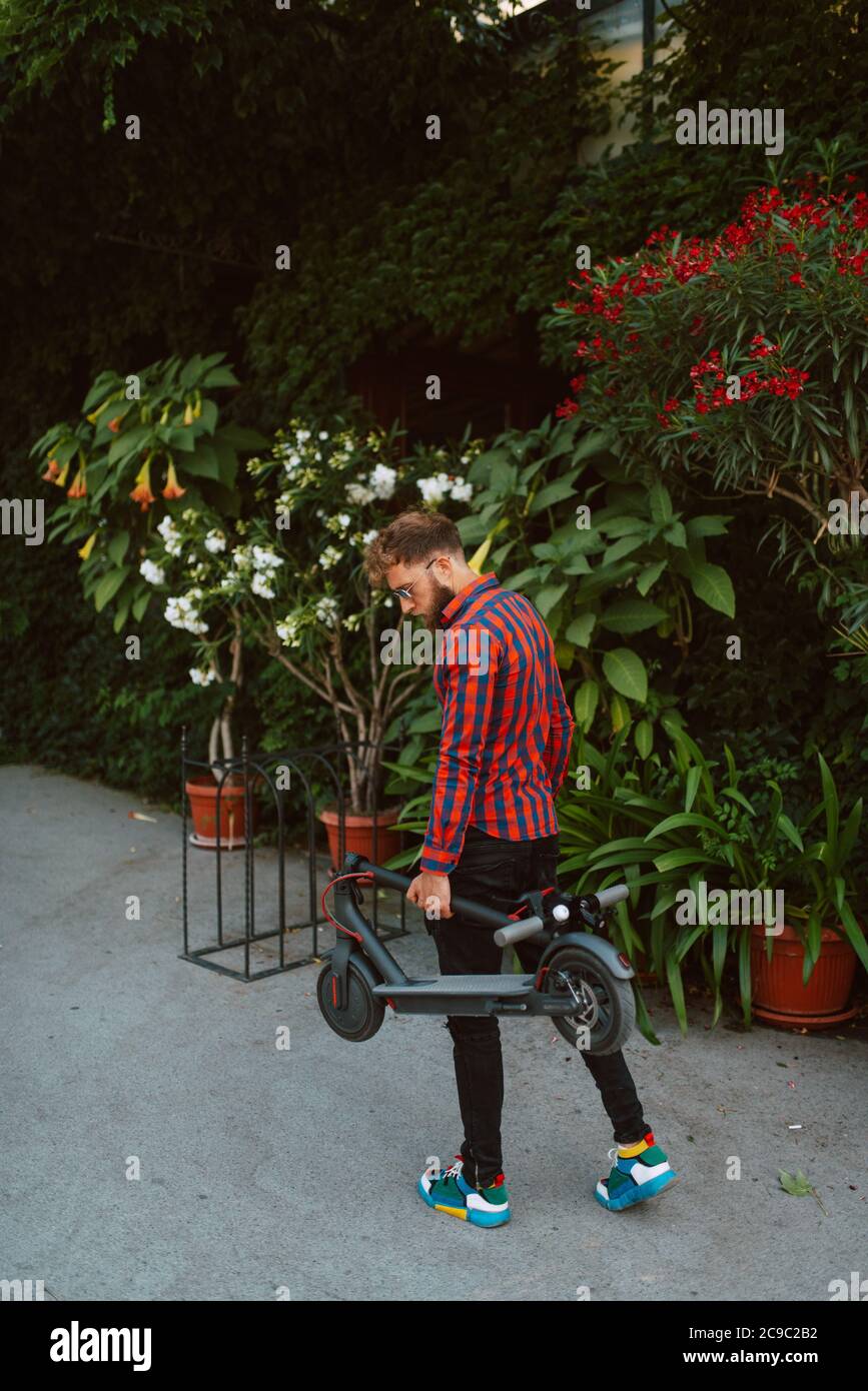 Ein hübscher junger bärtiger Kaukasusmann mit Sonnenbrille steht Auf der Straße mit E-Scooter Stockfoto