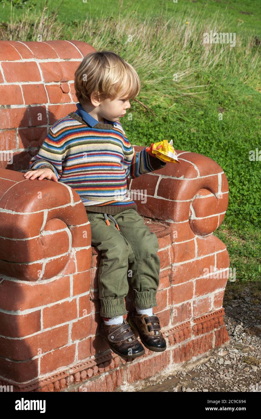 Kleiner Junge (2/3 Jahre) sitzt auf 'Se Bryck', einer Skulptur von Amy Cooper, Tremenheere Scupture Gardens, Penzance, Cornwall, Großbritannien. MODELL FREIGEGEBEN Stockfoto