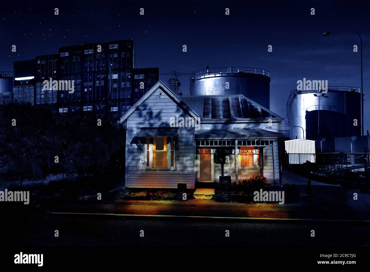 Ein Holzhaus aus dem frühen 20. Jahrhundert, umgeben von einem kommerziellen Containerhafen und großen Öltanks. Nachts aufgenommen, mit Sternen und Fensterbeleuchtung. Stockfoto