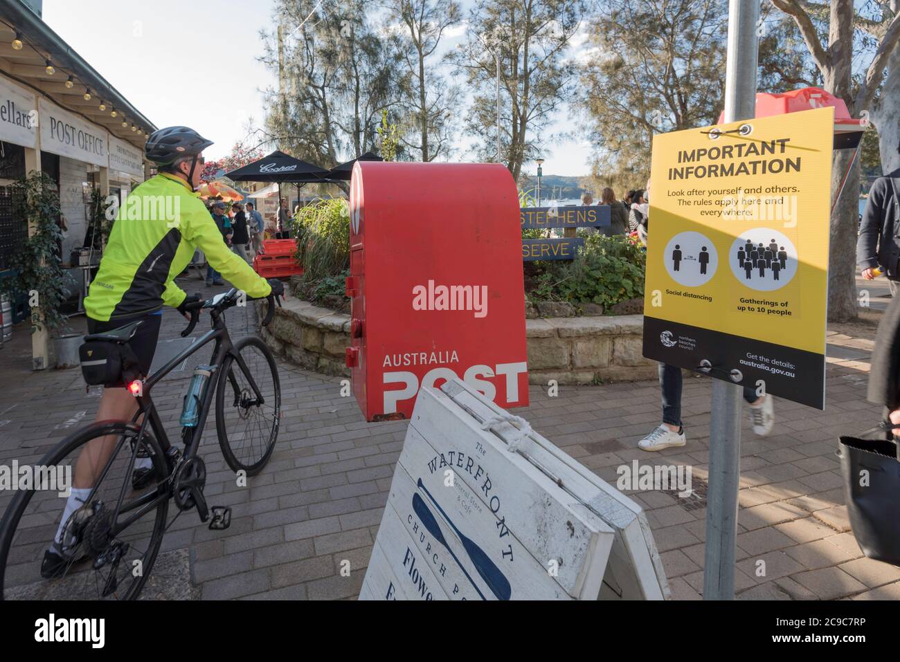 Eine Person, die eine leuchtend gelbe Hi-vis Jacke trägt, geht mit dem Fahrrad an einem Covid-19 (Coronavirus) sozialen Distanzierungsschild am Church Point plaza vorbei. Stockfoto