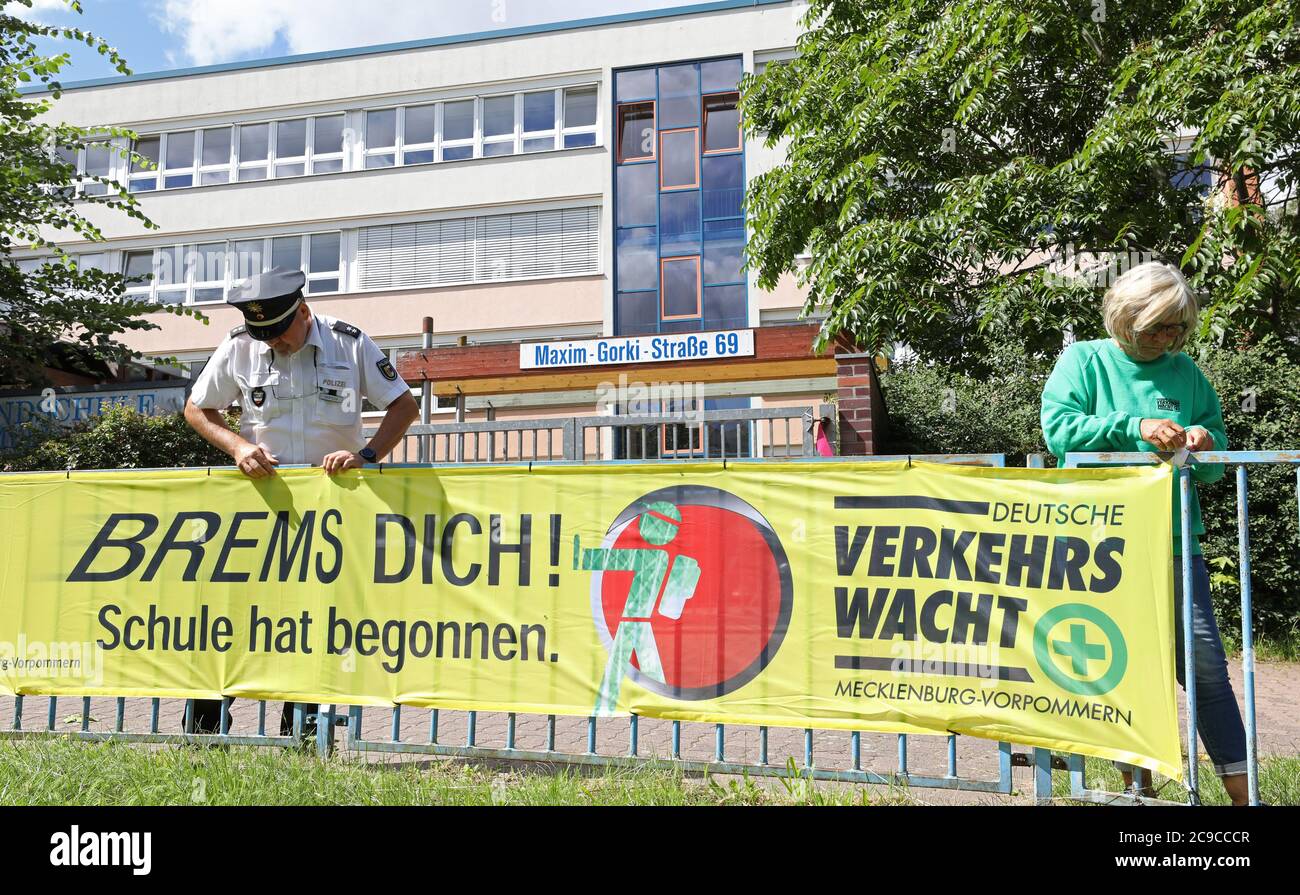 Rostock, Deutschland. Juli 2020. Matthias Bartsch, Präventionsbeauftragter der Rostocker Polizei, und Angelika Stiemer, Verkehrswache, befestigen vor der Grundschule am Mühlenteich ein Transparent mit der Aufschrift "Brems dich! Die Schule hat begonnen." Am 03.08.2020 beginnt wieder der Unterricht für rund 150,000 Schüler in Mecklenburg-Vorpommern. Quelle: Bernd Wüstneck/dpa-Zentralbild/ZB/dpa/Alamy Live News Stockfoto