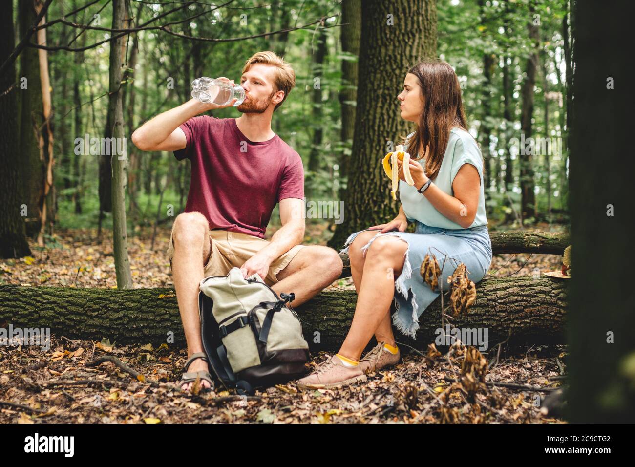 Die Leute essen Banane und trinken Wasser aus der Plastikflasche, während sie auf Holz sitzen. Paar Wanderer machen Pause für Essen und Trinken im Wald auf gefallen Stockfoto