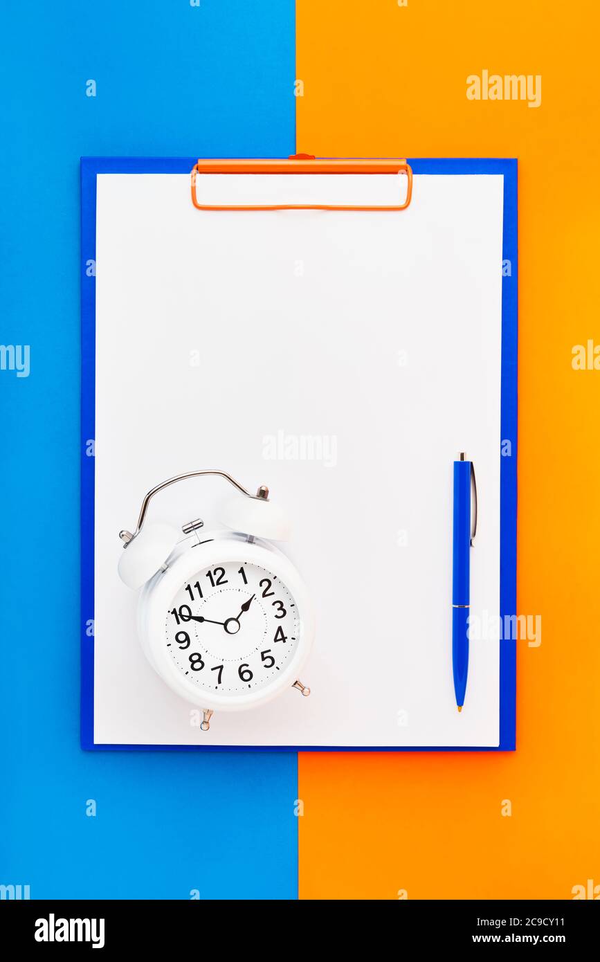 Leeres Clipboard-Modell, ein blauer Stift und ein weißer Wecker auf dem blau-orangefarbenen Hintergrund. Draufsicht, Kopierbereich für Text. Zurück in die Schule, Deadline Stockfoto