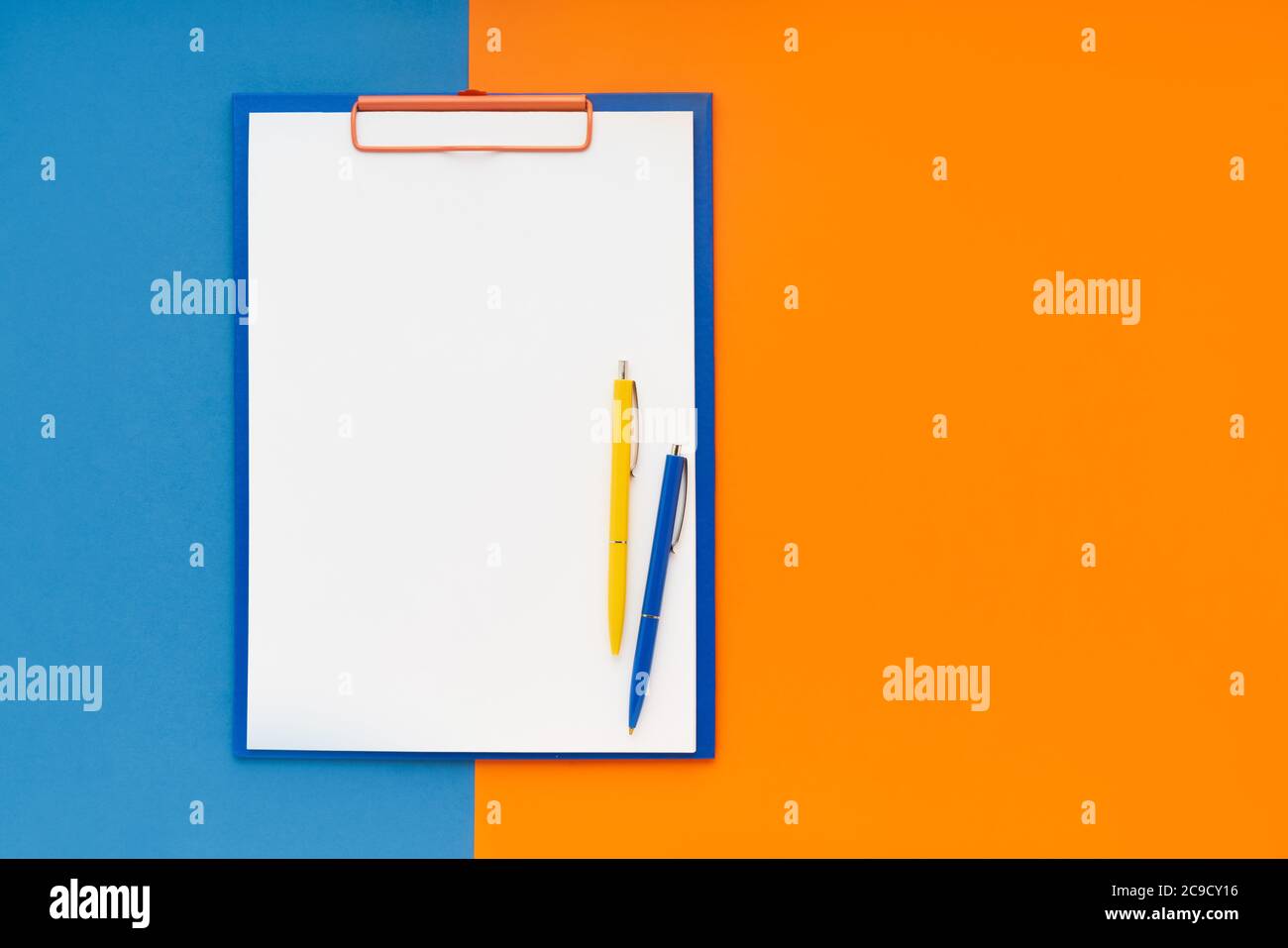 Leeres Clipboard-Mockup und zwei Stifte auf blau-orangefarbenem Hintergrund. Draufsicht, Kopierbereich für Text. Zurück zur Schule, Deadline, Morgenkonzept. Stockfoto