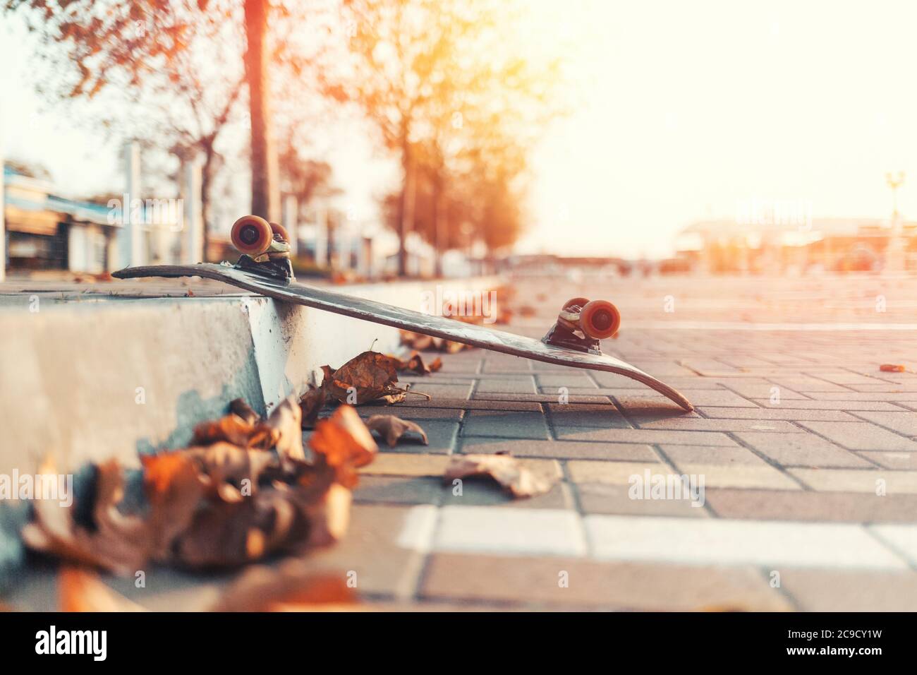 Skateboarding. Ein umgedrehtes Skateboard liegt auf dem Bürgersteig, mit heruntergefallenen Blättern. Im Hintergrund eine leere Straße der Stadt. Sonnenlicht. Speicherplatz kopieren Stockfoto