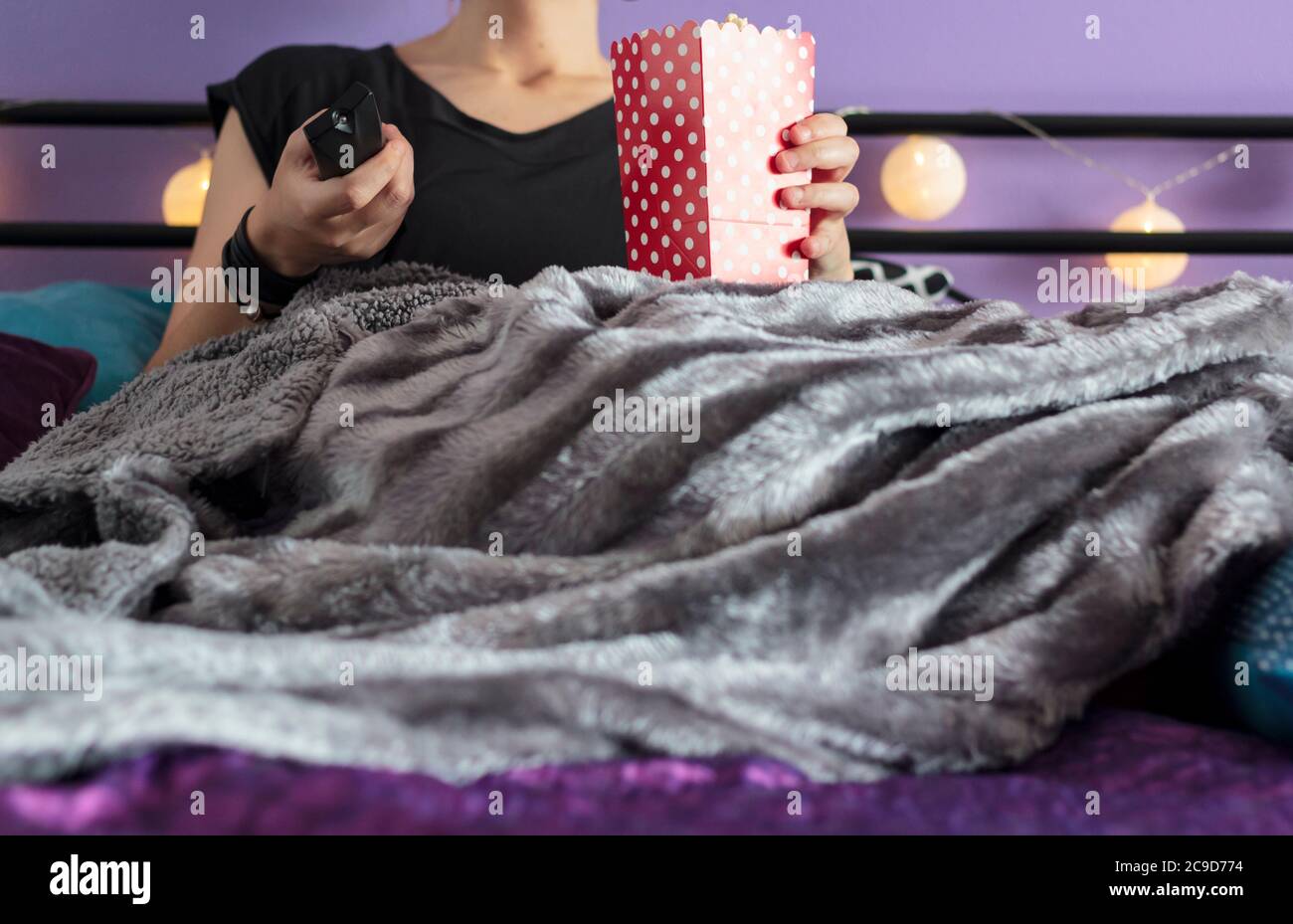Frau im Bett mit Decke bedeckt, die Popcorn und Fernbedienung hält Stockfoto